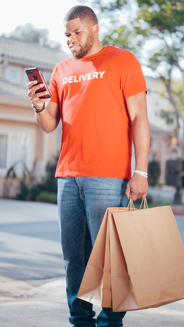 Man in a delivery shirt texting while holding paper packages on a sunny street.